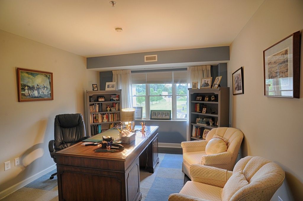 A cozy home office with a large desk, comfortable armchairs, and bookshelves under natural light from a window.