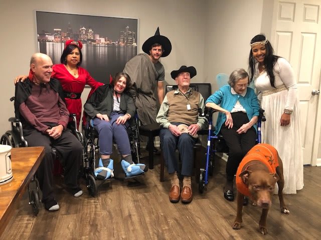 A group of people in costumes gather indoors, with some seated in wheelchairs and a dog in the foreground.