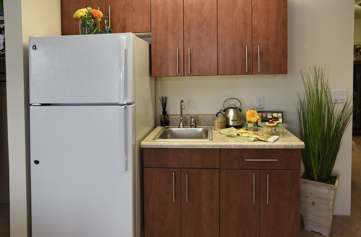 A small kitchen area with wooden cabinets, a refrigerator, and a countertop featuring a sink, kettle, and flowers.