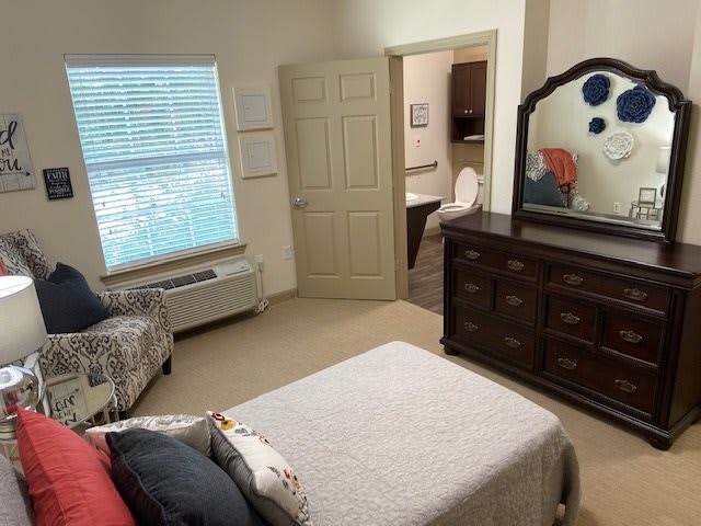 A neatly arranged bedroom with a bed, dresser with mirror, and a view of an adjoining bathroom.