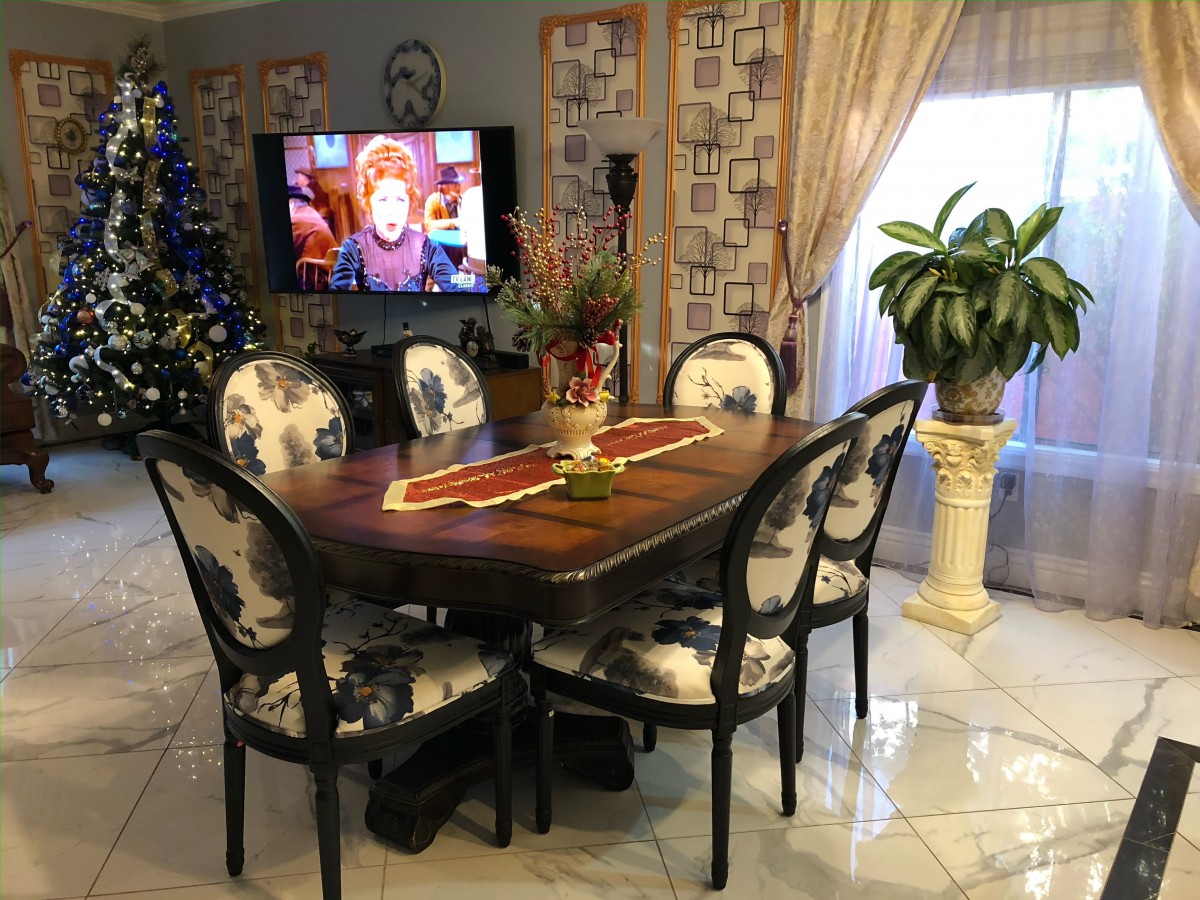 A cozy dining room with a decorated table, surrounded by floral-patterned chairs, and a television displaying a movie. A Christmas tree adds a festive touch to the space.