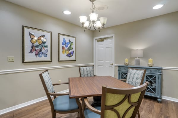 A cozy dining room featuring a wooden table, upholstered chairs, and butterfly-themed artwork on the walls. A chandelier hangs above, adding to the warm ambiance.