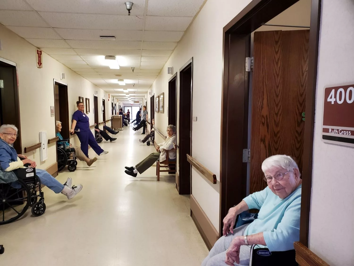 Elderly residents in wheelchairs line the hallway of a nursing home, accompanied by caretakers.