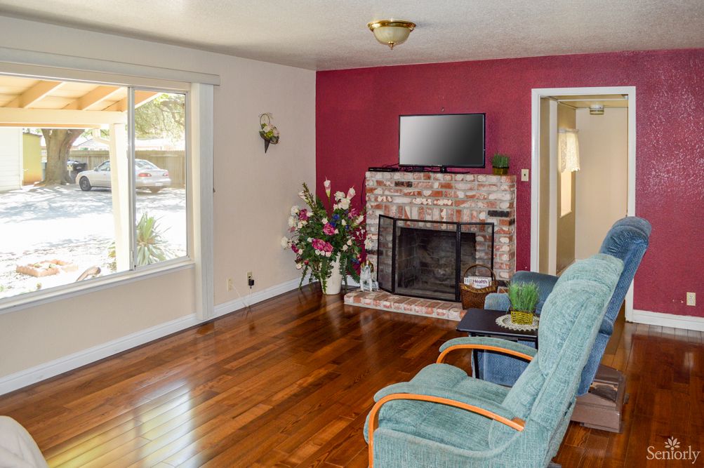 A cozy living room with hardwood floors, a brick fireplace, and a television mounted above. The room features a red accent wall, large window, armchair, and decorative plant.