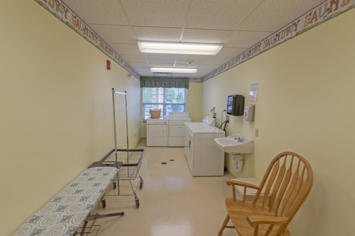 A laundry room with washing machines, a dryer, ironing board, and a chair.