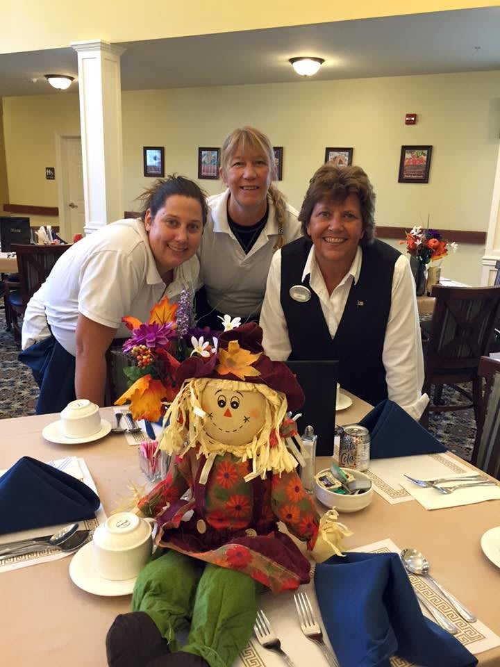 Three people posing with a scarecrow decoration on a table in a restaurant setting.