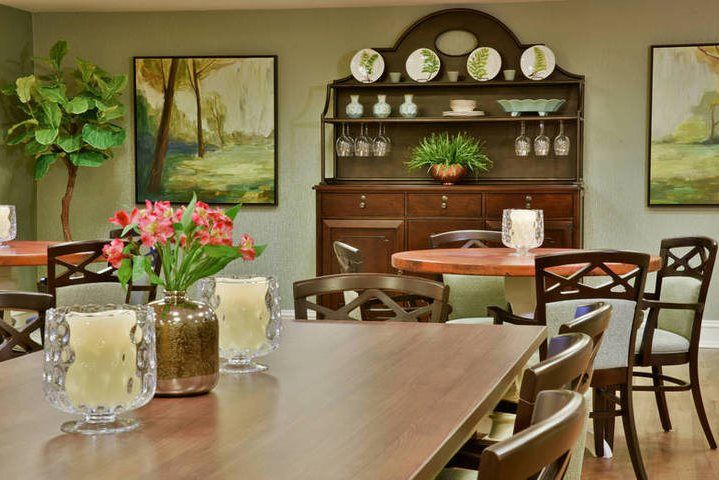 A cozy dining room with a wooden table, chairs, and a vintage cabinet adorned with decorative plates. The space is brightened by flowers and artwork on the wall.