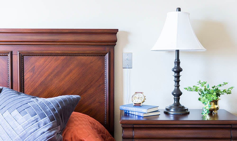 A cozy bedroom setup featuring a wooden headboard, bedside table with a lamp, alarm clock, and stacked books.