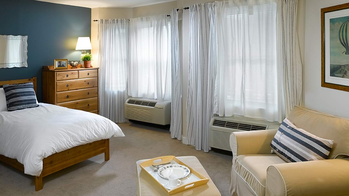 A cozy bedroom featuring large windows with light curtains, a wooden dresser, and comfortable seating.