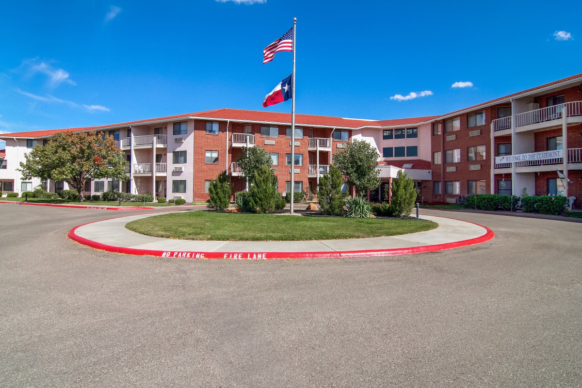 A three-story apartment complex with American and Texas flags flying in a well-landscaped roundabout on a sunny day.