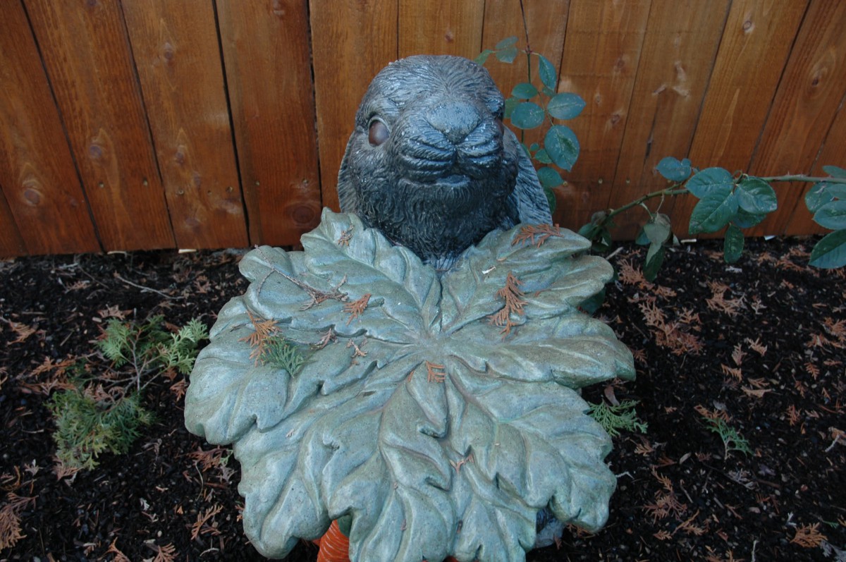 A stone owl sculpture partially hidden behind large leaf-shaped decoration in a garden.