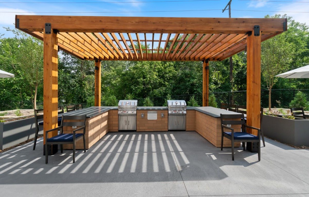 An outdoor kitchen with a pergola, featuring barbecue grills and seating on a sunny patio.