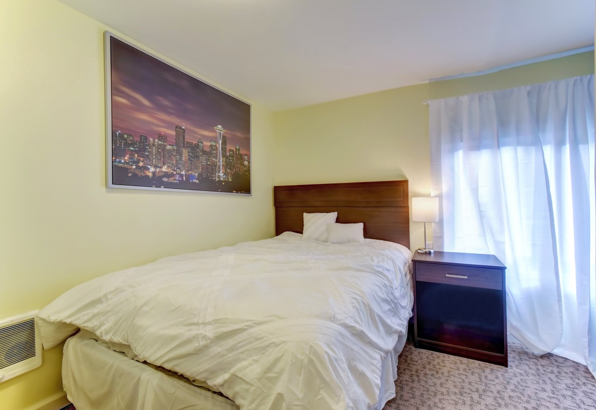 A cozy bedroom with a neatly made bed, nightstand, and a large cityscape painting on the wall.