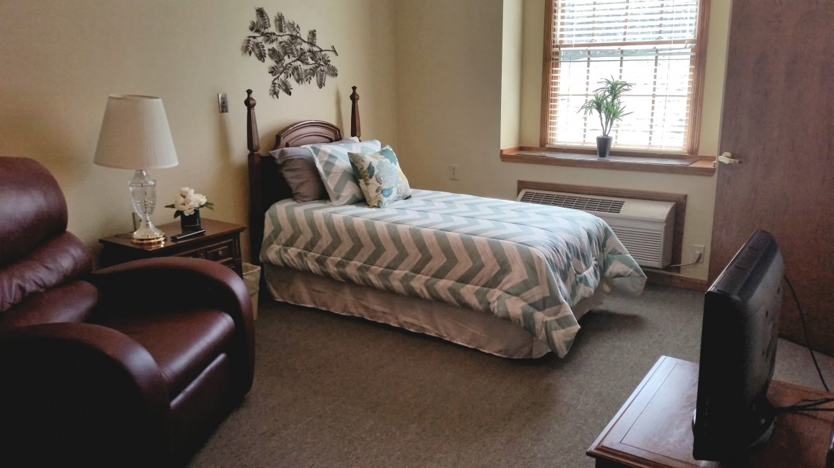 A cozy bedroom with a single bed, decorated with cushions and a patterned duvet, featuring a comfortable chair and a window letting in natural light.