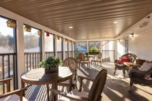 A cozy screened porch with wooden furniture and potted plants, bathed in sunlight.