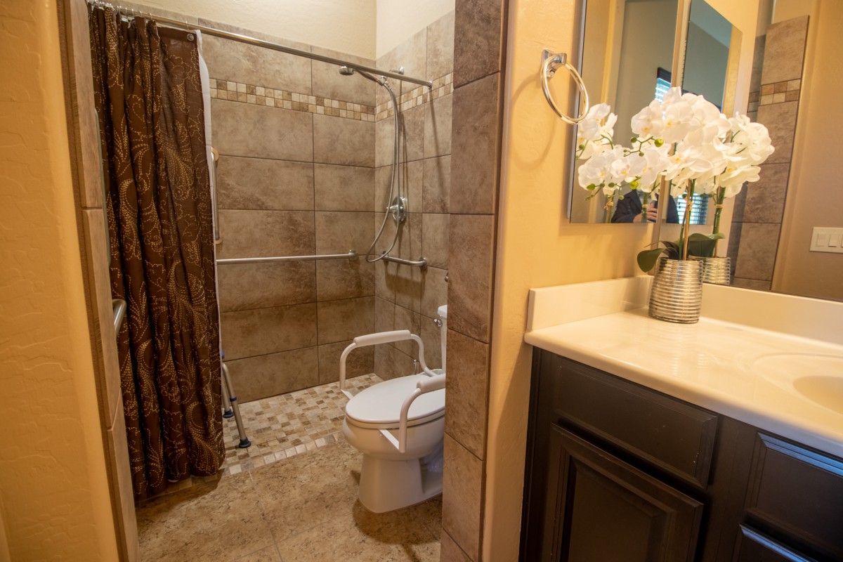 A bathroom with a shower, toilet equipped with handrails, sink, and decorative flowers.