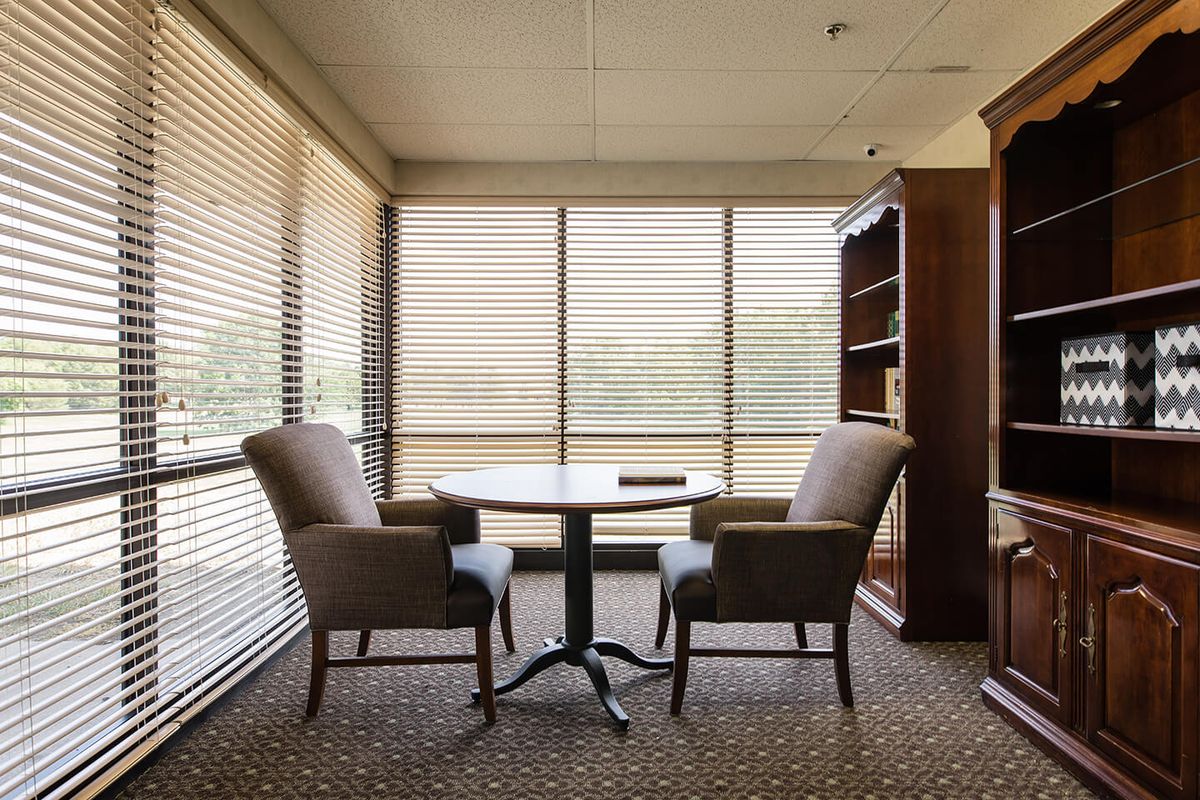 A quiet meeting room with two chairs and a table by large windows with blinds.