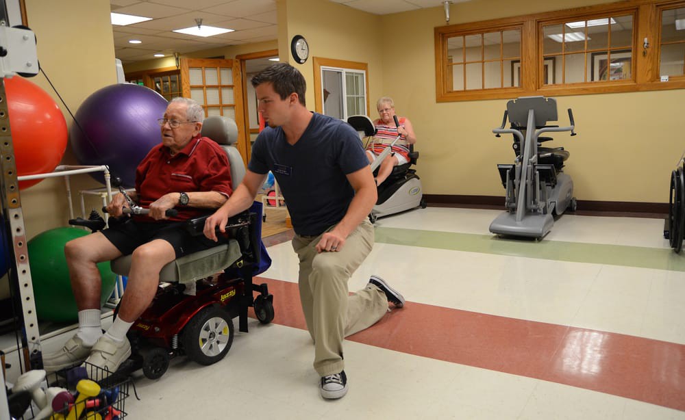 A healthcare professional assists a senior in a wheelchair with physical exercises in a rehabilitation center.
