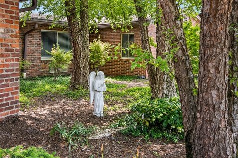 A serene garden with an angel statue surrounded by trees in front of a brick house.