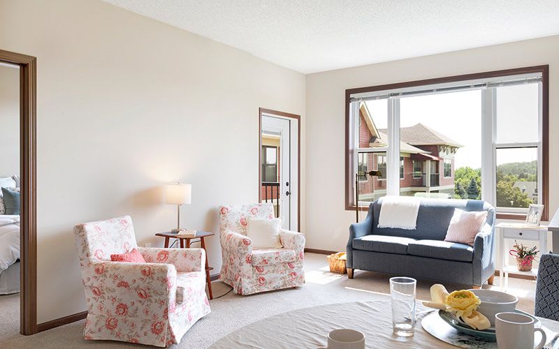 A cozy living room featuring floral armchairs, a dark blue sofa, and a large window letting in natural light.
