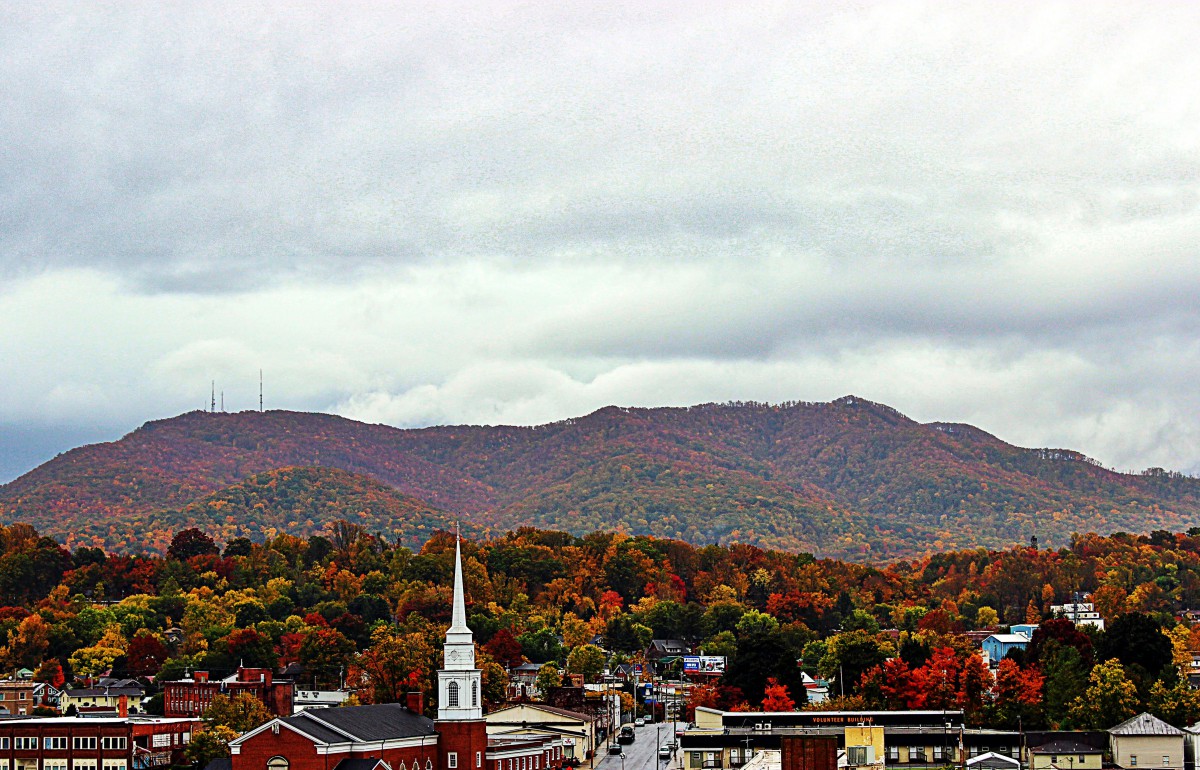 A scenic view of a town surrounded by colorful autumn foliage with mountains in the background under a cloudy sky.