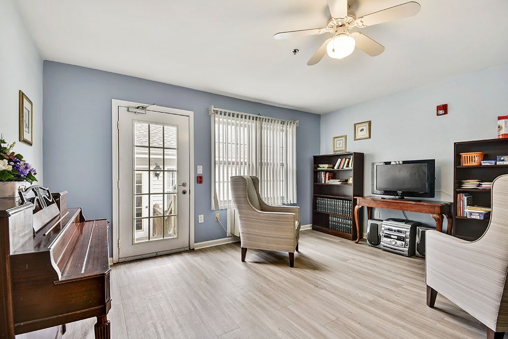 A cozy living room with blue walls, featuring an armchair, television, and bookshelf. The space is well-lit with natural light from large windows and a glass door.