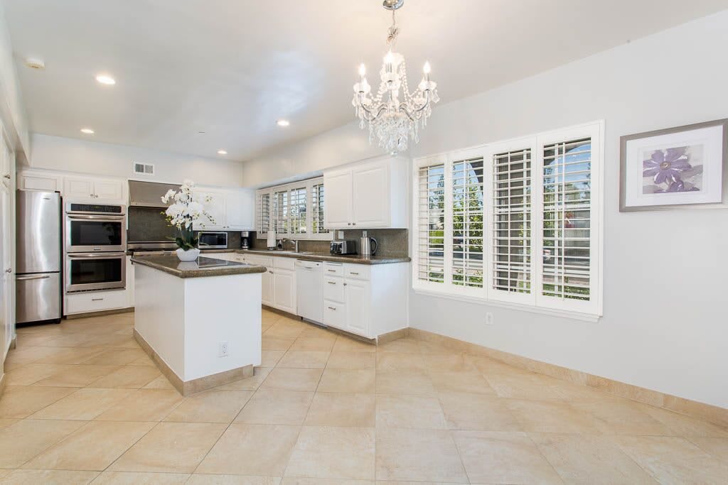 A modern kitchen with white cabinets, a central island, and a chandelier, featuring tiled floors and bright lighting.