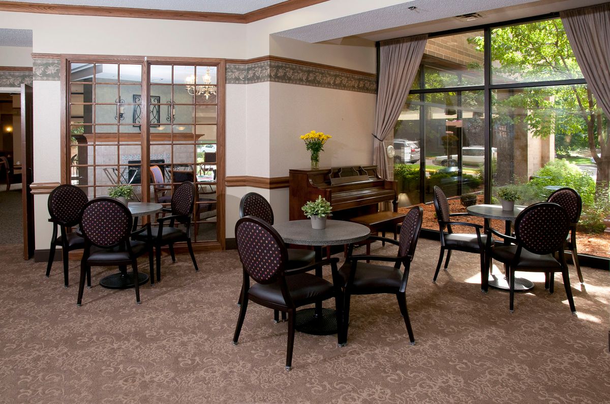A cozy seating area with round tables and upholstered chairs, featuring a piano and large windows with drapes.