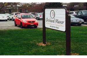 A parking lot with a sign for Orchard Manor and a red car parked nearby.
