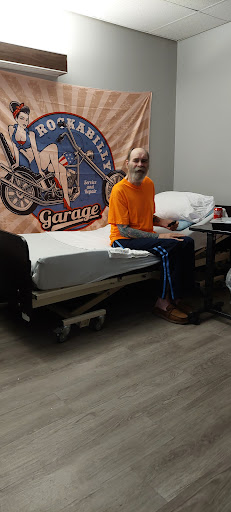A patient sitting on a hospital bed, smiling in an orange shirt and blue pants.