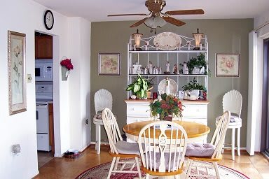 A cozy dining room with a round wooden table and white chairs, adorned with plants and a bookshelf in the background.