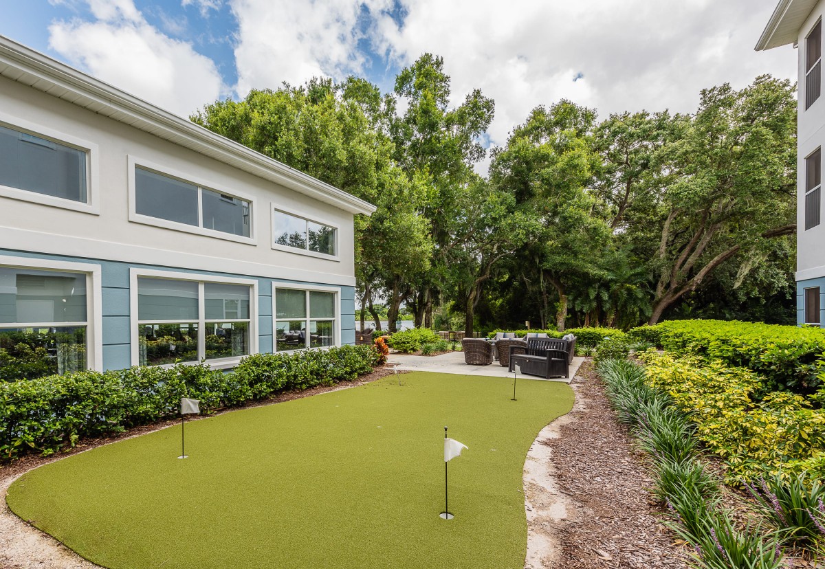 A backyard featuring a putting green and a patio area surrounded by lush greenery.