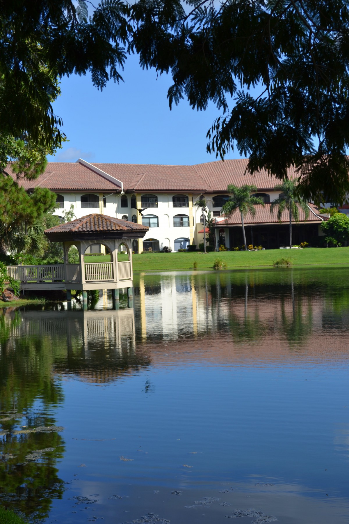 A serene landscape featuring a building with a red-tiled roof, reflected in a calm lake alongside a gazebo and lush greenery.