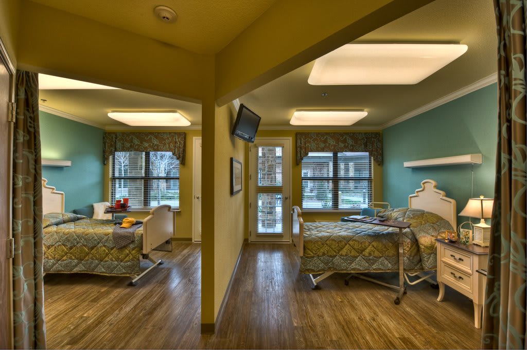 A hospital room with two patient beds, each having its own window and television.
