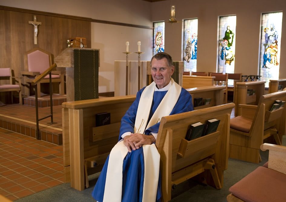 A priest seated in a church with stained glass windows in the background.