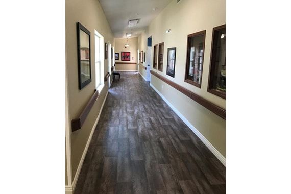 A long hallway with wooden flooring, decorated with artwork on the walls and illuminated by natural light through windows.