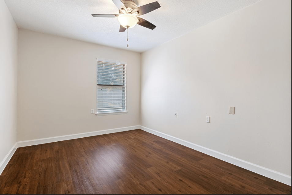 A spacious empty room with wood flooring, a ceiling fan, and a window.