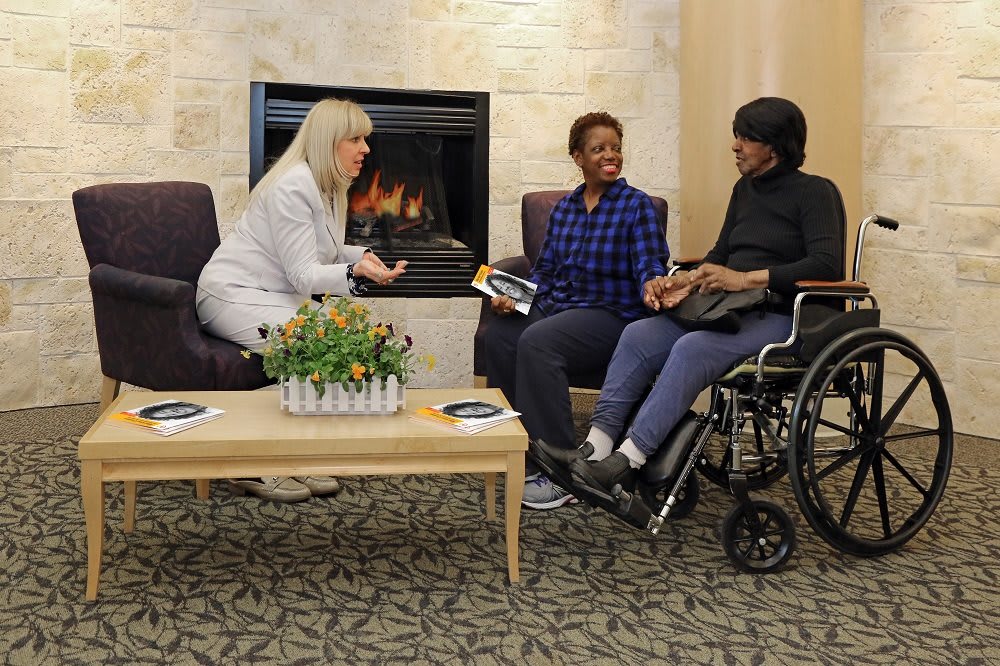 Three people having a conversation in a cozy indoor setting by the fireplace, one of them in a wheelchair.