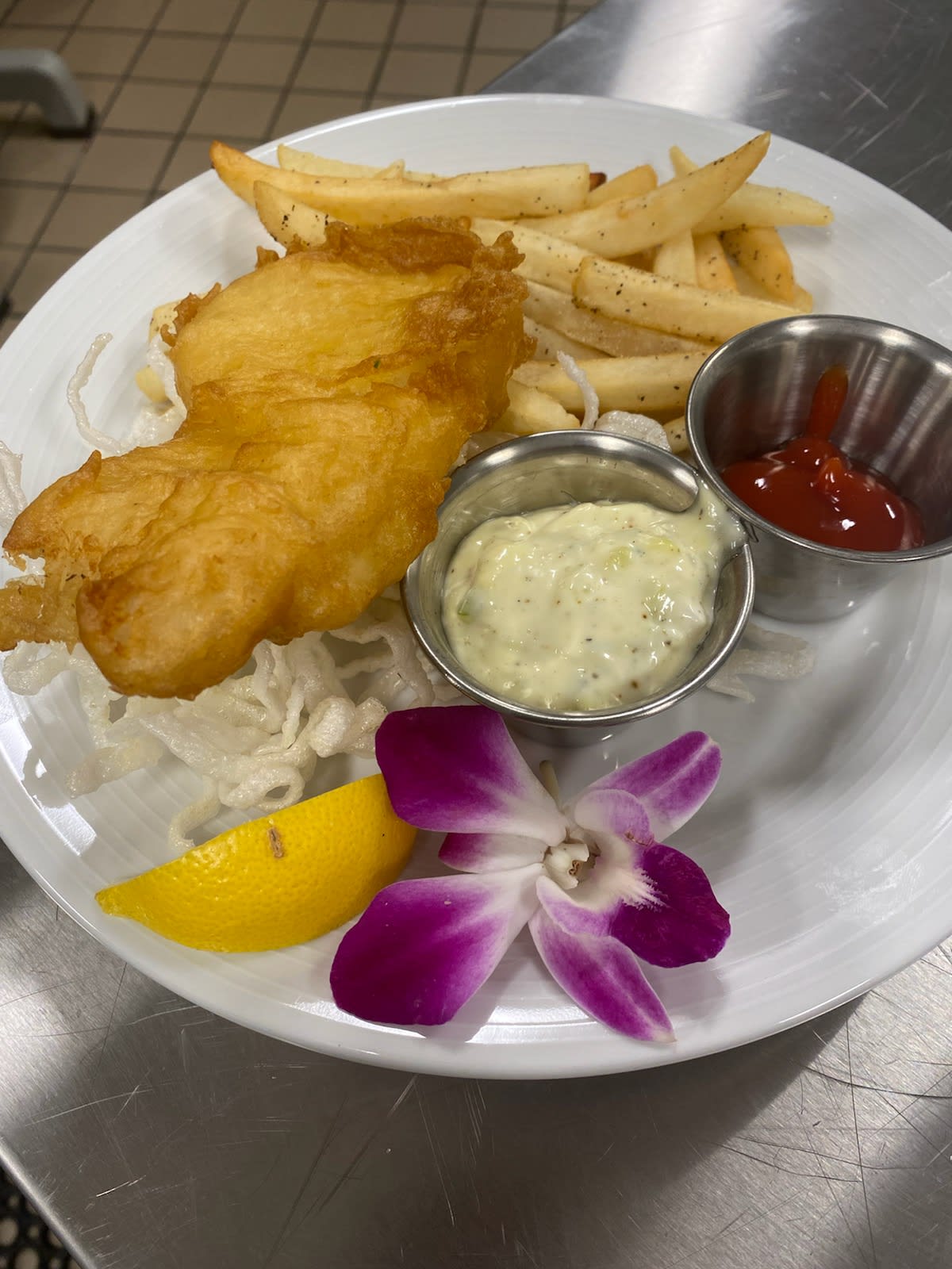 A plate of fish and chips with tartar sauce, ketchup, a lemon wedge, and a flower garnish.