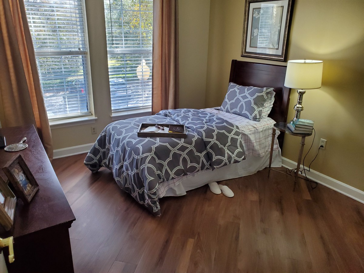 A cozy bedroom with a single bed, patterned bedding, and natural light streaming through large windows.