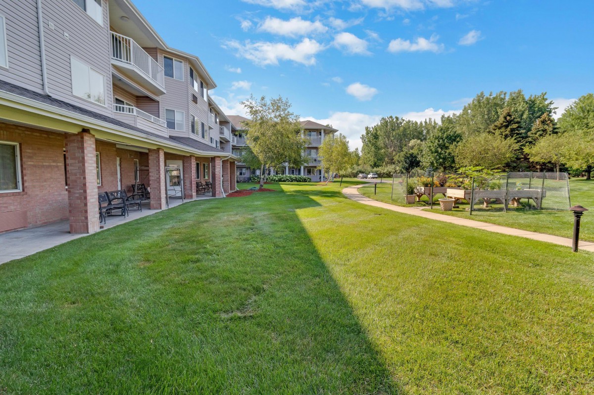 A view of an apartment complex with a well-maintained lawn and pathway, surrounded by trees under a clear blue sky.