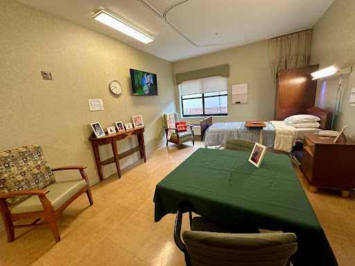 A well-lit hospital room featuring a bed, a table with chairs, and personal photographs.