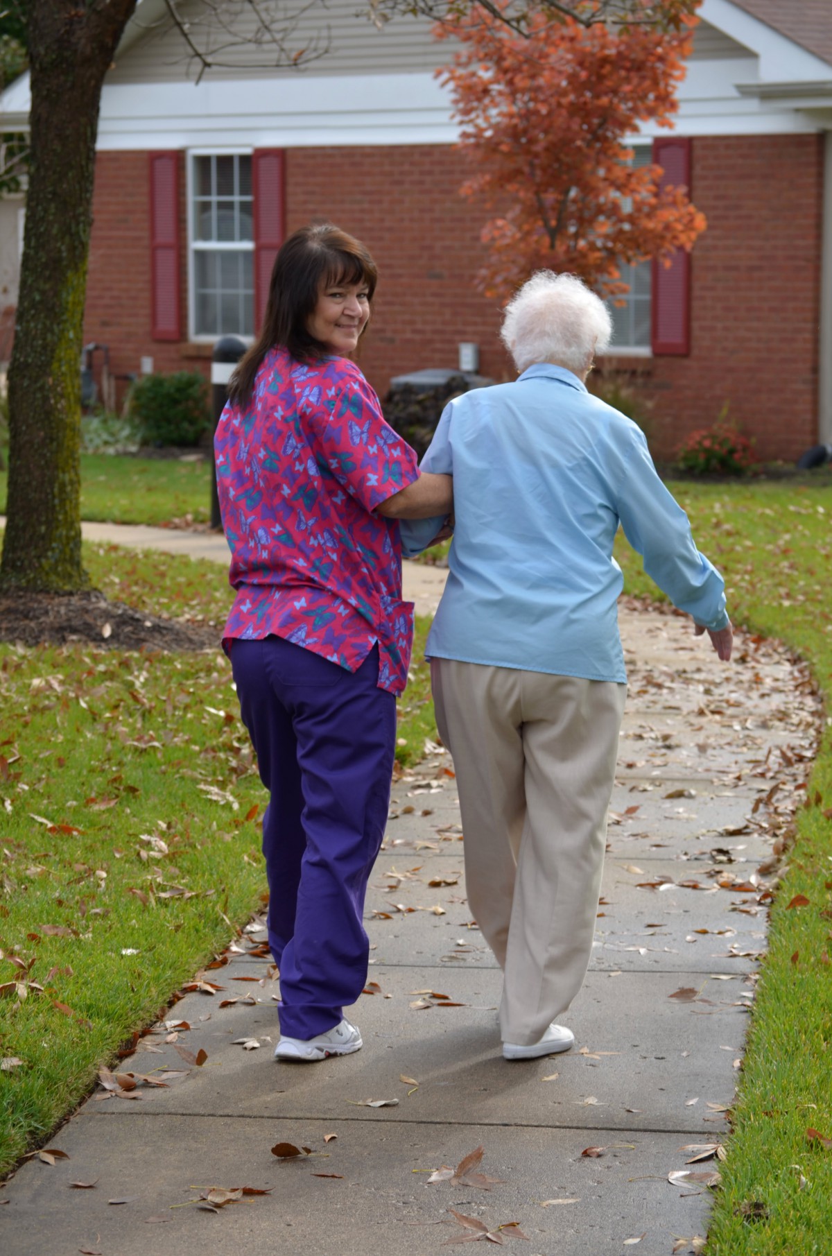A caregiver assists an elderly person during a walk on a leaf-strewn path.