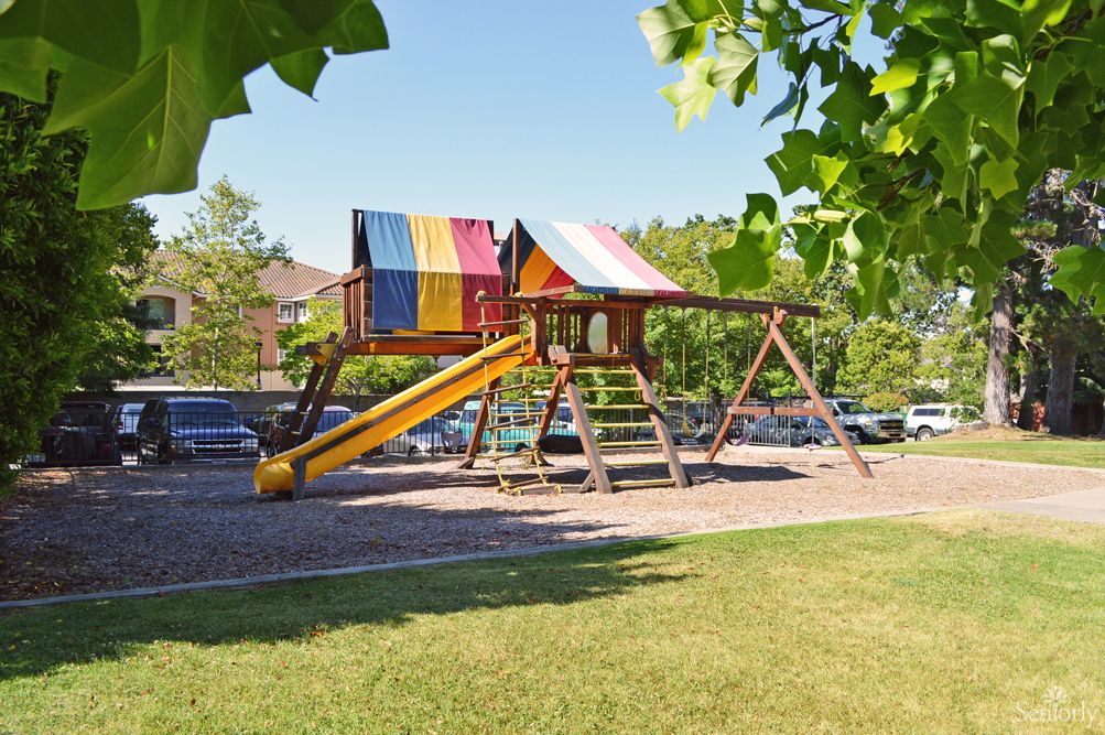 A colorful playground with a slide and swing set on a sunny day, surrounded by trees and grass.