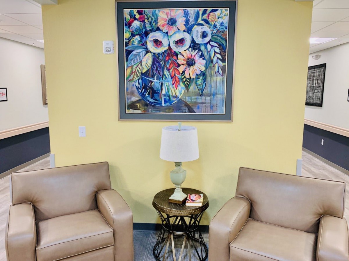 A cozy interior space featuring two armchairs, a side table with a lamp, and a vibrant floral painting on the wall.