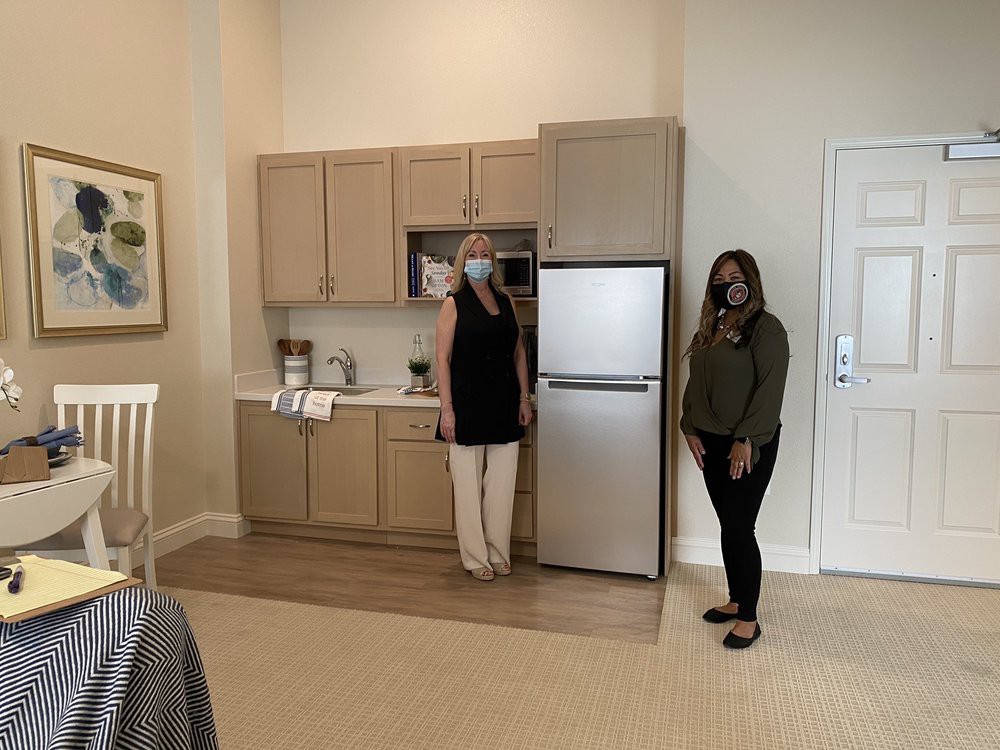 Two people standing in a small kitchen area with cabinets and a fridge.