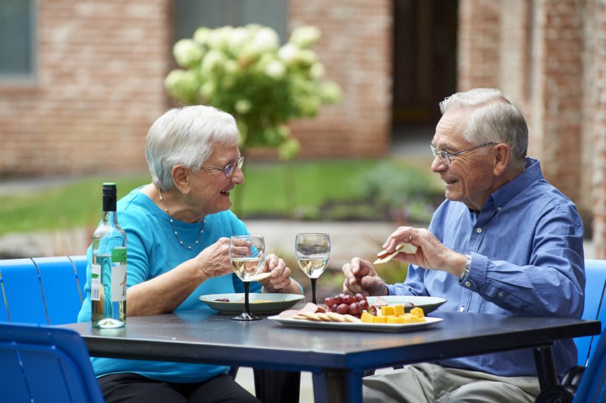 An elderly couple enjoys a meal together outside, sharing wine and conversation.