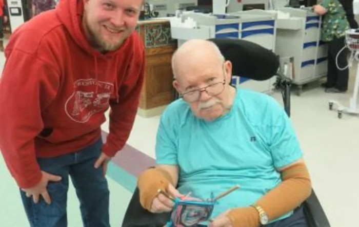 An elderly man in a wheelchair holding an item, with another man standing beside him.