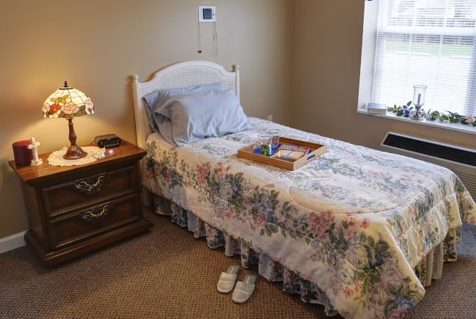 A cozy bedroom featuring a single bed with a floral comforter, accompanied by a nightstand and lamp. Slippers are placed neatly under the bed and a tray rests on top.