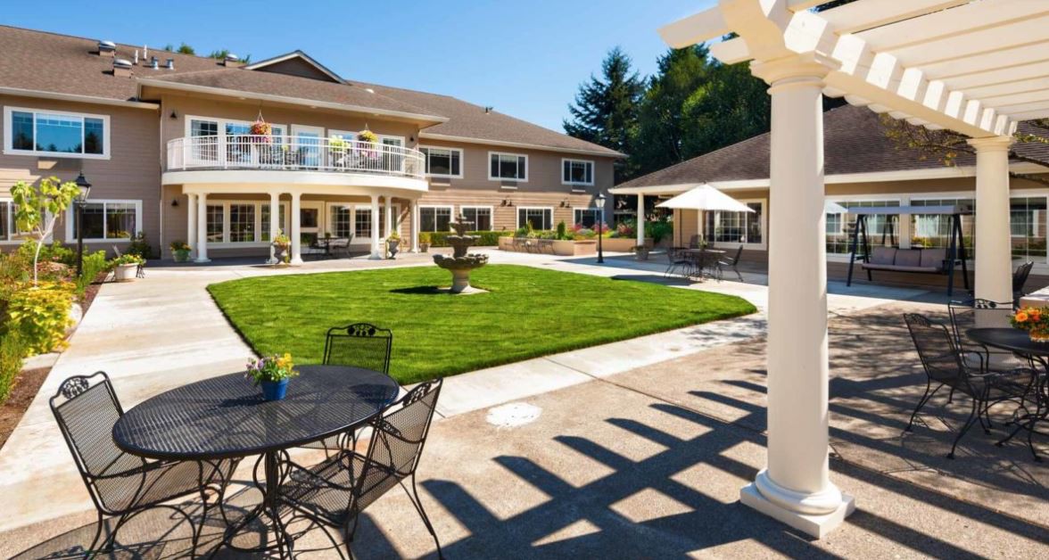 A sunny patio area with outdoor seating, surrounded by a well-maintained garden and a two-story building in the background.